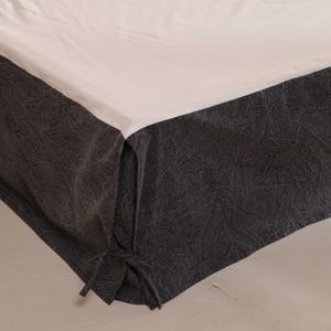 Può includere: Una gonna da letto con un tessuto grigio scuro strutturato e un lenzuolo superiore color crema. La gonna da letto ha un design a balze ed è legata agli angoli.