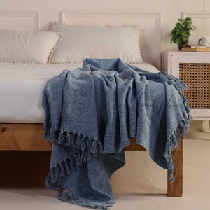 Könnte beinhalten: Eine blaue gewebte Decke mit Fransen, die über einer Holzbank drapiert ist. Die Decke hat ein strukturiertes Aussehen und ist einfarbig blau. Der Hintergrund umfasst ein Bett mit weißen Kissen und einen Nachttisch.