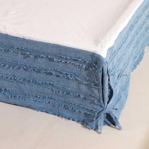 May include: A close-up of a bed with a white sheet and a blue bed skirt. The bed skirt has a fringed texture and is tied at the corners. The overall aesthetic is casual and inviting.