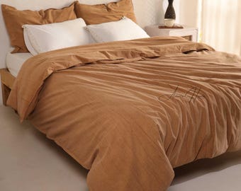 May include: A bed with a tan duvet cover and matching pillowcases. Two white pillows are visible. The bed has a wooden frame. The image is taken in a well-lit room.