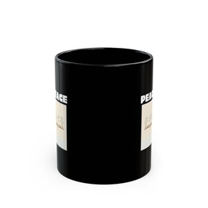 May include: A black ceramic mug featuring a rectangular graphic on the side. The graphic displays the word PEACE in bold white capital letters above an illustration of an open book with small tree branches growing out of the pages.