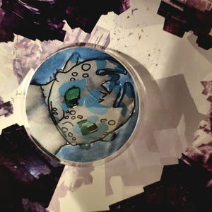 May include: A round, clear glass coaster with a watercolor illustration of a cartoon dragon's face. The dragon has green eyes and is set against a blue and white background. The coaster is surrounded by a purple and white abstract design.