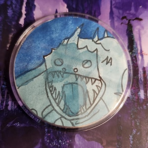 May include: A round, clear button with a watercolor illustration of a cartoon monster. The monster is primarily light blue with black outlines, featuring sharp teeth and a long tongue. The background is a gradient of blues.