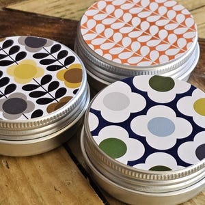 May include: Three small, round metal tins with patterned lids. The lids feature floral designs in various colours, including yellow, brown, and blue, set against white and navy backgrounds. The tins are arranged on a wooden surface.