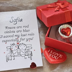 May include: A red gift box with a bow, open to reveal a heart-shaped candy. A card with the name "Sofia" and a poem is next to the box. A small red heart and a heart-shaped "I Love You" patch are also present.