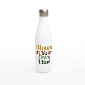 Puede incluir: Botella de agua aislada blanca con tapa plateada. La botella presenta el texto "Bloom at Your Own Time" en letras doradas, verdes y marrones. La botella está diseñada para mantener las bebidas frías o calientes.