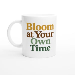 Puede incluir: Taza de cerámica blanca con el texto "Bloom at Your Own Time" en amarillo mostaza, marrón y verde. La taza tiene un asa curva y está sobre un fondo blanco.