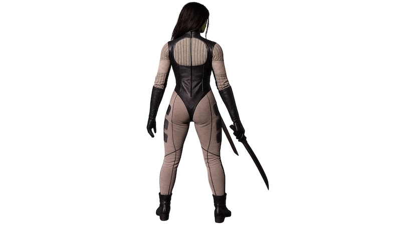 May include: A figure in a black and tan bodysuit with black gloves and boots, holding a black sword. The bodysuit has a high collar and a leather-like appearance. The figure is viewed from the back against a white background.