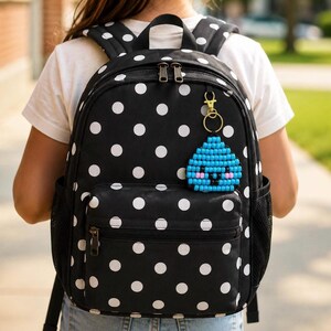 Puede incluir: Una mochila negra con estampado de lunares blancos que incluye un llavero dorado con forma de gota de agua azul. La gota está hecha de pequeñas cuentas y muestra una cara sonriente con mejillas rosadas, ojos y boca de color negro.