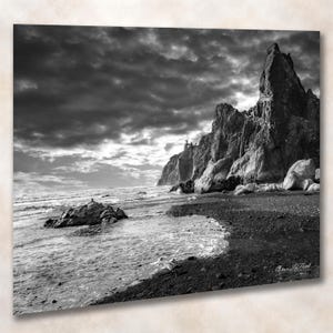 May include: A black and white landscape photograph featuring a rugged coastline with large, jagged rock formations rising from a dark, pebbled beach. Waves wash onto the shore under a dramatic, cloudy sky. The signature Vince LaPoint is visible.