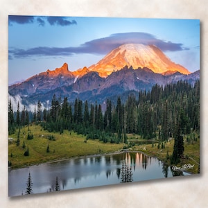 May include: A landscape photograph featuring a snow-capped mountain peak illuminated by golden sunlight, topped with a lenticular cloud. A dense forest of evergreen trees sits below the mountain, bordering a calm lake reflecting the scenery.
