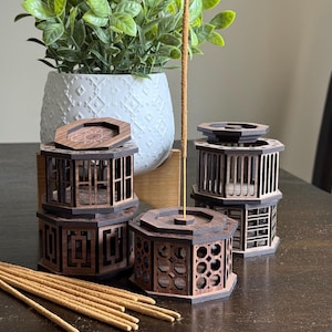 May include: Wooden incense holders in various sizes and designs. The holders are dark brown with intricate cut-out patterns. Incense sticks are visible, along with a white patterned vase with a green plant.
