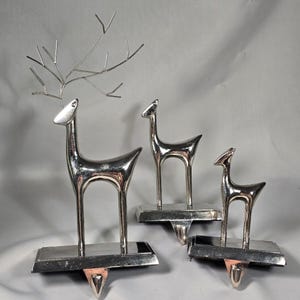 May include: A set of three silver-toned metal deer stocking holders. Each figure features a smooth, polished finish, standing on a rectangular base with a downward-pointing hook. The largest deer includes thin, wire-like antlers.
