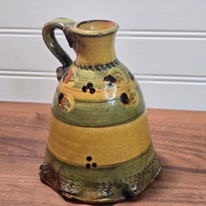 May include: A ceramic pitcher featuring a bell-shaped body with alternating horizontal bands of mustard yellow and olive green glaze. The surface is decorated with small dark brown floral motifs and a textured zigzag pattern along the base rim.