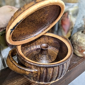 Puede incluir: Una cazuela de cerámica marrón hecha a mano con tapa. La cazuela tiene un exterior acanalado, un asa y una característica central en forma de cono. La tapa está parcialmente abierta, revelando el interior. La cazuela tiene una estética rústica y terrosa.