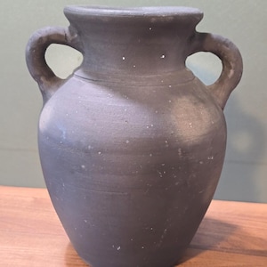 May include: A dark gray, matte-finished ceramic vase with a rounded body, a wide neck, and two symmetrical handles on either side. The surface features subtle horizontal ridges and small, light-colored speckles across the exterior.