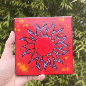 May include: Square red ceramic tile featuring a stylized sun design with a central red circle and blue outlined rays. The tile has a textured surface with yellow accents and black outlines.