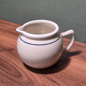 May include: A small, off-white ceramic creamer pitcher with a rounded body, a single handle, and a thin, dark blue horizontal line encircling the upper portion of the vessel. The pitcher features a small, integrated pouring spout.
