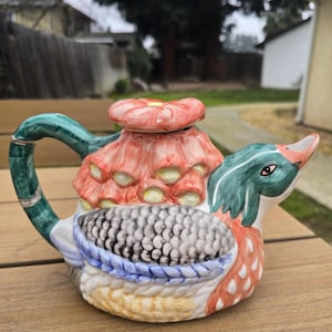 May include: A ceramic teapot shaped like a duck, featuring a green handle and beak, with a red and orange floral design on the lid and body. The teapot has a detailed, textured appearance with blue and white accents.