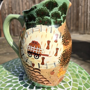 May include: A hand-painted ceramic pitcher with a farm scene. The pitcher features a light green handle and rim, with a detailed illustration of a farm with a wagon, fence, and trees. The background is a mix of tan, brown, and green.