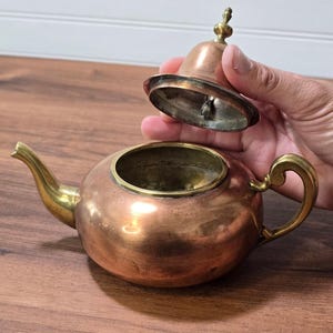 May include: A copper teapot with a brass spout, handle, and lid finial. The teapot has a rounded body and a wide opening. The lid is held separately above the opening, revealing a brass rim and a metallic interior finish on the lid's underside.