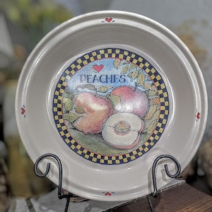 May include: A white ceramic bowl with a peach design, featuring the word "PEACHES" and a heart. The bowl is supported by a black metal stand. The design includes peaches, leaves, and a checkered border.