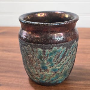 May include: A small ceramic cup with a smooth, dark metallic brown rim and a textured, mottled body featuring shades of deep teal and black. The vessel has a rounded shape with a slightly flared opening and a rough, uneven surface finish.