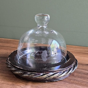 May include: A clear glass cloche with etched floral designs sits atop a dark brown, round wooden base with a braided edge. The cloche has a small knob on top. The base is approximately 6 inches in diameter.