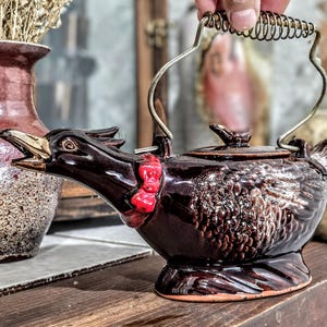 May include: A brown ceramic teapot shaped like a rooster, with a red bow tie and gold accents on the beak. The teapot has a metal handle with a coiled grip. A vase with dried flowers is in the background.