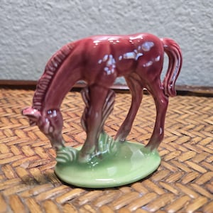 Puede incluir: Figurita de cerámica de un caballo burdeos en posición de pastoreo. El caballo tiene un acabado brillante y se apoya sobre una base ovalada verde con hierba estilizada. La figura es un objeto decorativo, probablemente para exhibición. Medidas en centímetros.