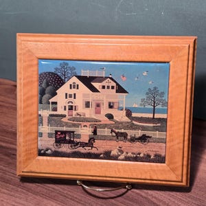 Könnte beinhalten: Eine rechteckige Keramikfliese mit einer Volkskunstszene eines weißen Hauses am Meer, gerahmt in hellem Holz. Das Bild zeigt eine Pferdekutsche mit der Aufschrift Betsy's, einen weißen Lattenzaun, eine kleine amerikanische Flagge und einen blauen Himmel.