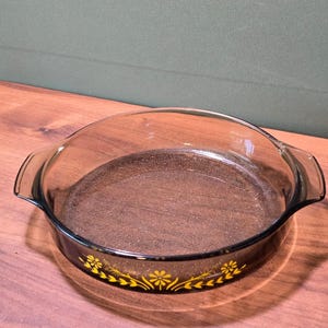 Peut inclure: Un plat de cuisson rond en verre brun foncé transparent doté de deux poignées latérales intégrées. L'extérieur du plat présente une bande décorative composée de motifs floraux et de feuilles jaunes entourant la base du récipient en verre.
