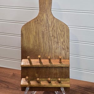 May include: A vertical wooden wall-mounted rack shaped like a cutting board with a handle at the top. It features two horizontal shelves, each equipped with four small wooden pegs for hanging items. The wood has a medium brown finish with visible grain.