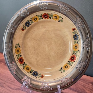 May include: A round ceramic plate with a cream-colored center featuring a fine crackle glaze and a floral border of red, yellow, and blue flowers. The plate has a wide, metallic silver rim decorated with embossed floral patterns around the edge.