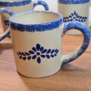 May include: A set of ceramic mugs with a cream-colored base and a blue speckled rim. Each mug features a blue floral motif on the front and a matching blue speckled handle. The design is consistent across the mugs, showcasing a simple, functional shape.