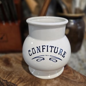 May include: A white ceramic jar with a rounded body and a flared rim. The word "CONFITURE" is written in dark blue script across the front, with decorative flourishes beneath. The jar sits on a wooden surface, suggesting a kitchen or dining setting.