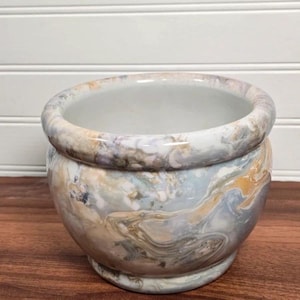 May include: A round ceramic planter featuring a marbled finish with swirling patterns of soft blue, cream, tan, and grey tones. The bowl-shaped vessel has a smooth, glossy surface, a thick rounded rim, and a wide opening, showcasing a white interior.