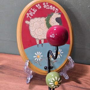 May include: An oval wooden wall hook featuring a painted sheep with a green wreath and the text Talk is Sheep. The design includes a red background, a blue foreground with white daisies, and two black metal hooks with a red ball and a green floral ball.