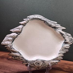 May include: A ceramic bowl with a smooth, glossy off-white interior and a textured, dark-veined exterior that mimics the appearance of weathered driftwood or tree bark, featuring irregular, jagged edges and a prominent knot detail at the base.