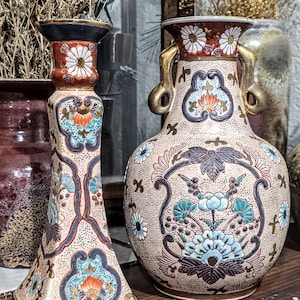 May include: Two antique ceramic vases with floral designs. The vase on the left is a candlestick holder with a square base, and the vase on the right is a round vase with gold handles. Both vases have a beige background with blue, orange, and white floral patterns.