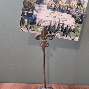 May include: A metal stand with a fleur-de-lis top and a leaf-shaped base holding a postcard of the California State Capitol building in Sacramento, featuring the text Sacramento California and State Capitol Building on the postcard.