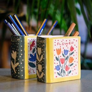 Può includere: Due portapenne in ceramica a forma di libro, uno verde scuro con un motivo floreale e la scritta "BLOOM", l'altro giallo con un motivo floreale e la scritta "FLOWER". Entrambi sono pieni di penne e matite.