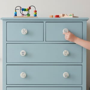 Peut inclure: Une commode bleu clair à cinq tiroirs avec des boutons blancs ornés d'animaux. Une main d'enfant atteint l'un des boutons. Un labyrinthe de perles en bois et des blocs de bois sont posés sur la commode.