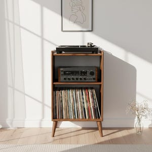 Può includere: Un supporto in legno per giradischi con tre ripiani. Il ripiano superiore contiene un giradischi nero. Il ripiano centrale ha un ricevitore stereo. Il ripiano inferiore è pieno di dischi in vinile.