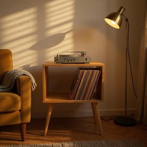 Può includere: Un tavolino in legno chiaro con un giradischi e una pila di dischi in vinile. Una lampada da terra dorata illumina la scena, accanto a una poltrona color senape. La stanza è immersa in una luce calda.
