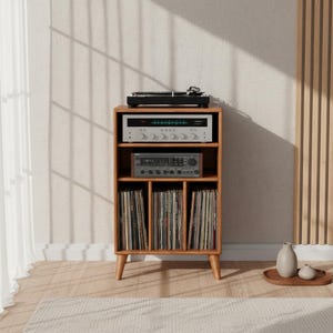 Könnte beinhalten: Ein Holz-Plattenspieler-Ständer im Mid-Century-Modern-Stil mit Plattenspieler, Stereo-Receiver und Verstärker. Der Ständer hat drei Regale, wobei Schallplatten auf dem unteren Regal aufbewahrt werden. Der Ständer ist aus hellbraunem Holz gefertigt und hat abgewinkelte Beine.
