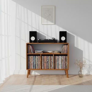 May include: A wooden record storage console with tapered legs, featuring an open top shelf for a turntable and two speakers, and three lower compartments filled with vinyl records. The unit has a warm brown finish and a rectangular frame.