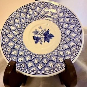 May include: A white and blue decorative plate with a floral design in the center. The plate has a blue geometric pattern around the rim. The plate is displayed on a brown stand.