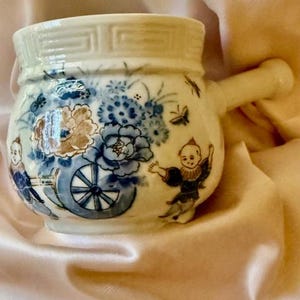 May include: A small, white ceramic soup bowl with a handle, decorated with blue floral and cart designs. The bowl features a decorative rim and illustrations of figures in traditional clothing. Butterflies are also depicted on the bowl.