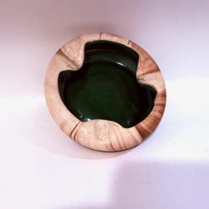 May include: A round ceramic ashtray with a dark green interior and a beige and brown marbled exterior. The ashtray has a unique, organic shape with a smooth, glossy finish. The rim has a textured, wood-like appearance.
